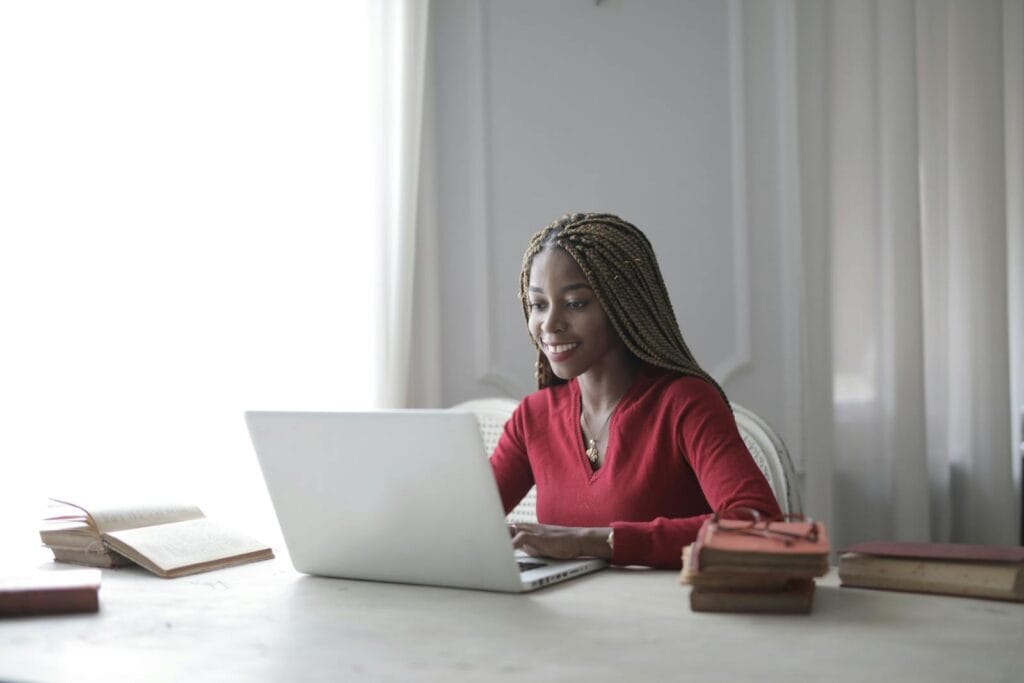 Vagas Remotas na Agência Mestre, salário de até R$ 5.100 Woman with braided hair smiling as she works on a laptop at home with books around her.