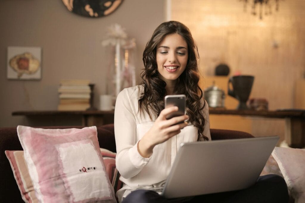 Assistente de SAC e mais Vagas Remotas Abertas! A woman enjoying leisure time using her smartphone and laptop in a cozy living room.
