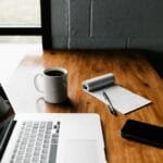 Assistente de Suporte ao Cliente Remoto, Oportunidade aberta! MacBook Pro, white ceramic mug,and black smartphone on table