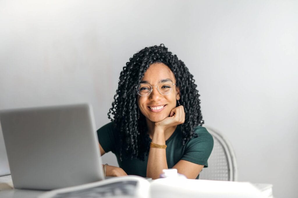 Aprendiz Administrativo Home Office Joyful businesswoman with curly hair smiling at camera while using laptop indoors.