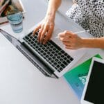 Assistente de Atendimento ao Cliente Remoto e mais Vagas A woman works on a laptop with a tablet in a modern home office setup.