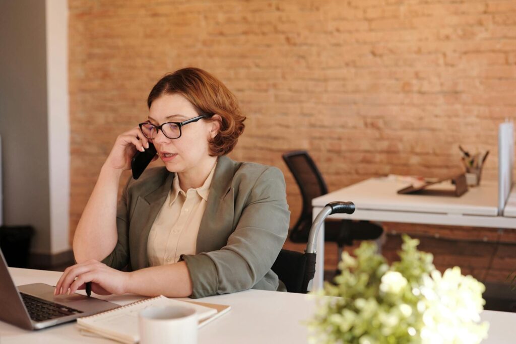 Vagas Remotas na Méliuz Professional woman in wheelchair working remotely on laptop and phone in a modern office.