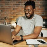 Vagas Remotas na ConteJá African American man smiling while working remotely on laptop from home office