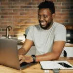Vagas Remotas na ConteJá African American man smiling while working remotely on laptop from home office