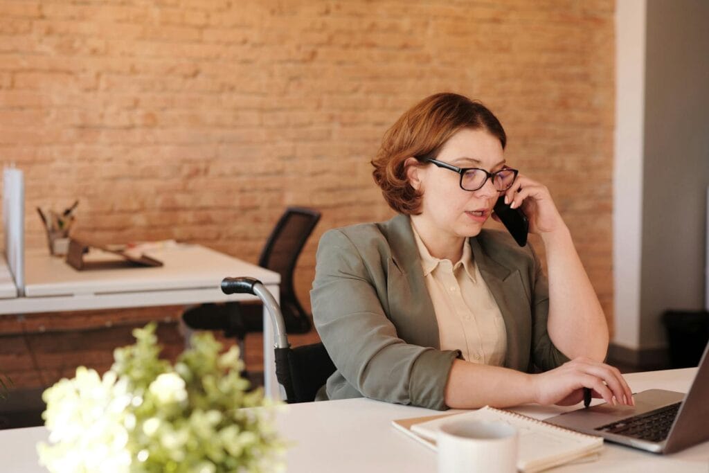 Vagas Remotas para Atendimento ao Cliente Professional woman in wheelchair working remotely on laptop and phone in a modern office.