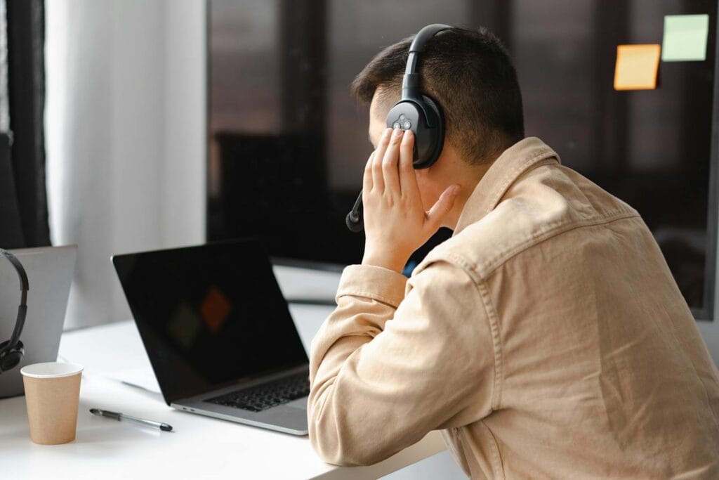 Trabalhe em Casa: 150 vagas na área de Atendimento A man wearing headphones works at a laptop in a modern office setting.