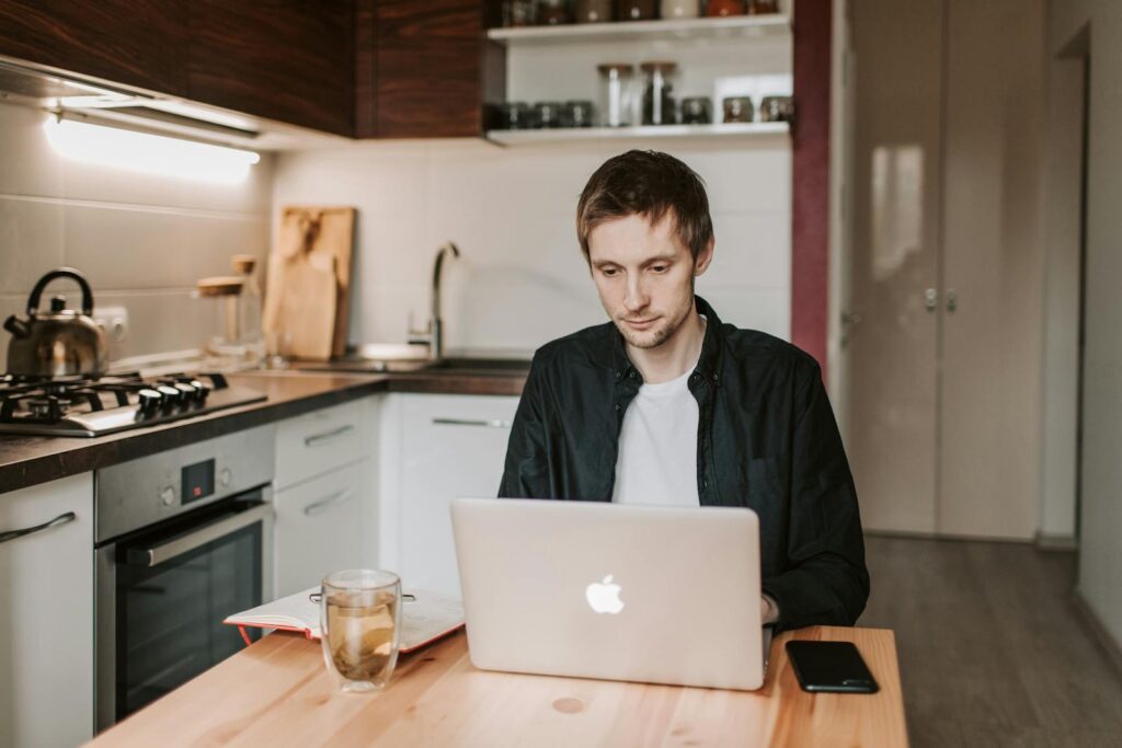 Remotas na Full Sales System Pensive young male freelancer in casual clothes watching netbook while sitting at wooden table with glass of water and copybook in apartment