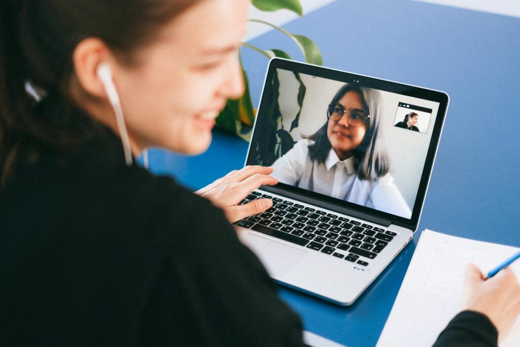 11 Vagas Remotas na Unicred Woman having a video conference on a laptop, smiling and taking notes.