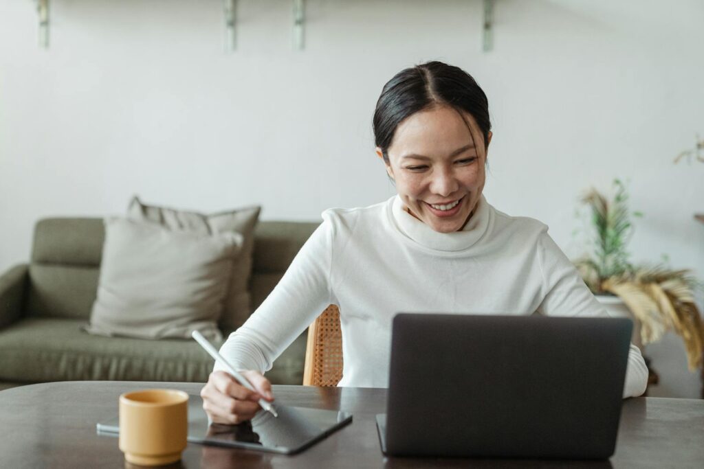 Auxiliar de Atendimento Home Office e Desenvolvedor BackEnd A cheerful woman uses a laptop and tablet for a video call, working remotely in a cozy living room.