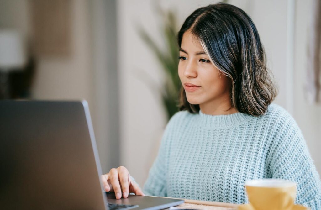Atendente Home Office e mais Vagas Remotas Confident young Latin American woman in casual clothes working remotely on netbook at table near mug with coffee in light apartment with plant with green leaves near wall