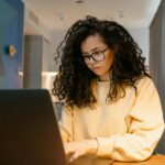 Assistente Administrativo sem experiência e mais! Woman in yellow sweater working on a laptop in a cozy home setting.