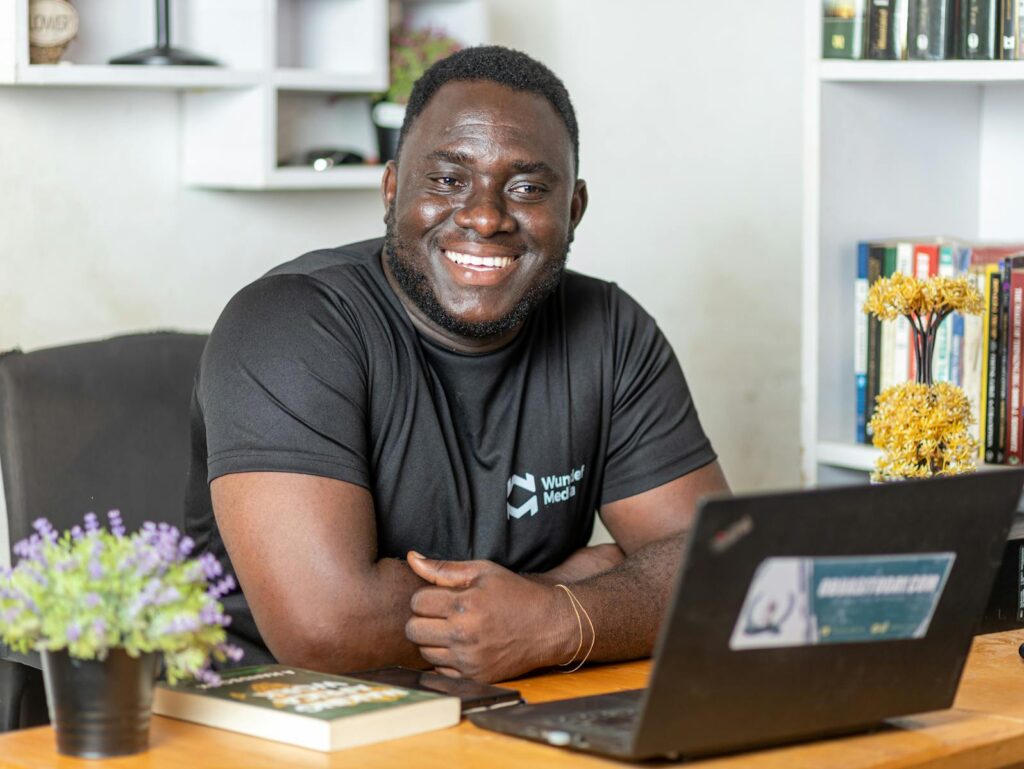 Analista de Suporte JR, Atendimento JR e Supervisor - Home Office A cheerful man working at his office desk using a laptop, surrounded by books and plants.