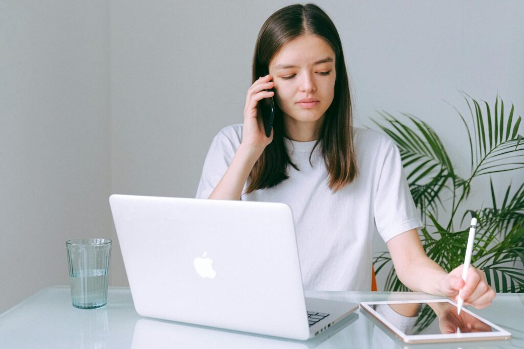 Analista de Suporte Home Office A young woman multitasking with a phone, laptop, and tablet in a bright home office setting.