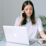 Analista de Suporte Home Office A young woman multitasking with a phone, laptop, and tablet in a bright home office setting.