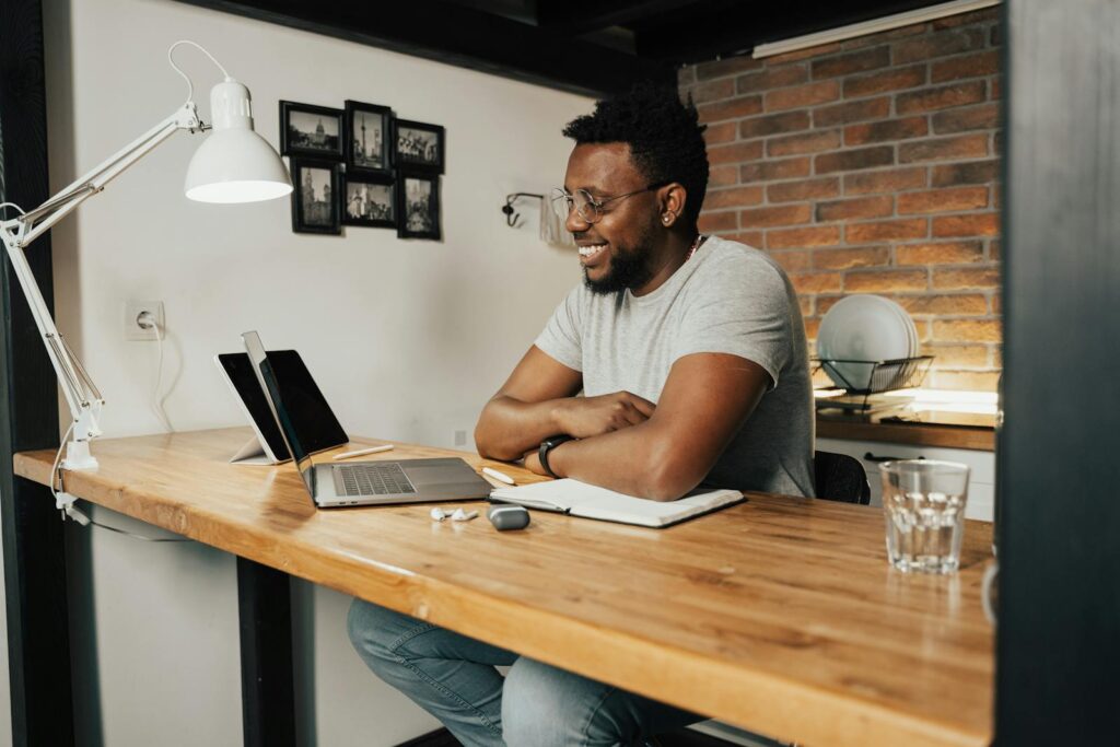 Vagas Remotas na Avanti African American man smiling and working remotely from home using a laptop and digital tablet.