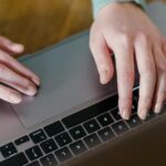 Vagas Remotas na Riachuelo, incluindo Assistente Administrativo From above of crop person using touchpad of laptop while browsing internet sitting near wooden table