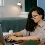 Emprego Remoto Sem Superior e salário de R$ 6.000 ou mais A woman focusing on remote work in a comfortable bedroom with modern decor and laptop.