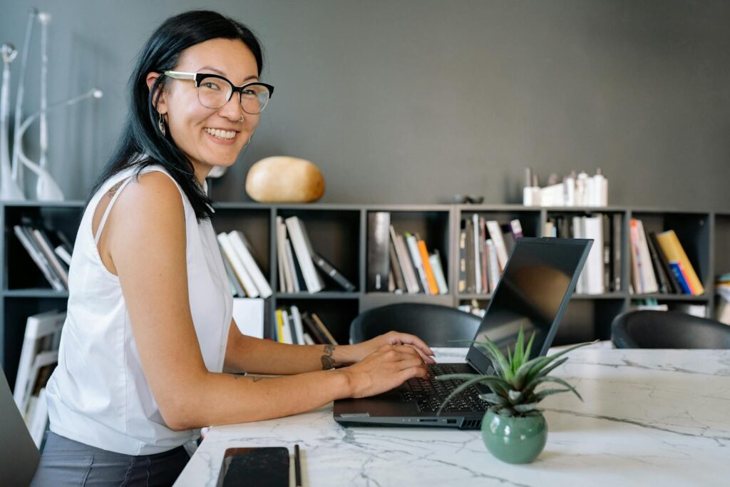 Vagas Remotas na Mercos Cheerful businesswoman using laptop at a desk in a stylish office setup with bookshelves.