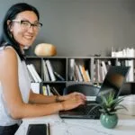 Vagas Remotas na Mercos Cheerful businesswoman using laptop at a desk in a stylish office setup with bookshelves.