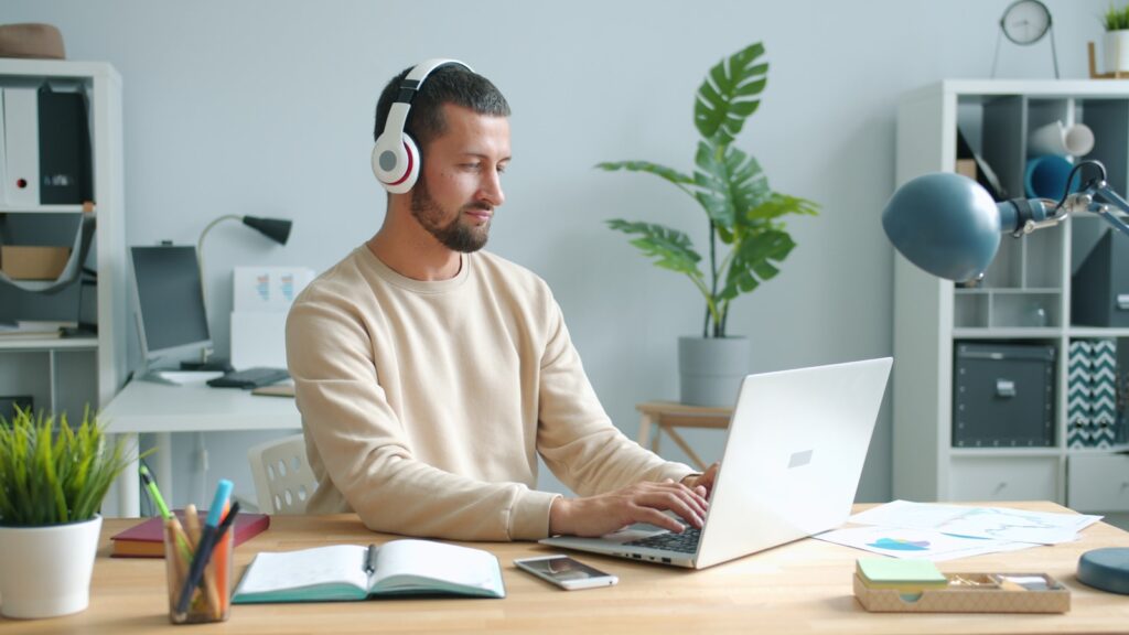 Vagas Remotas para Ensino Médio: 200 Oportunidades de Home Office Man wearing headphones works on laptop at desk.