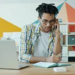 CLT Remoto com salário até R$ 10.000 Man talking on phone at desk with laptop and notebook.