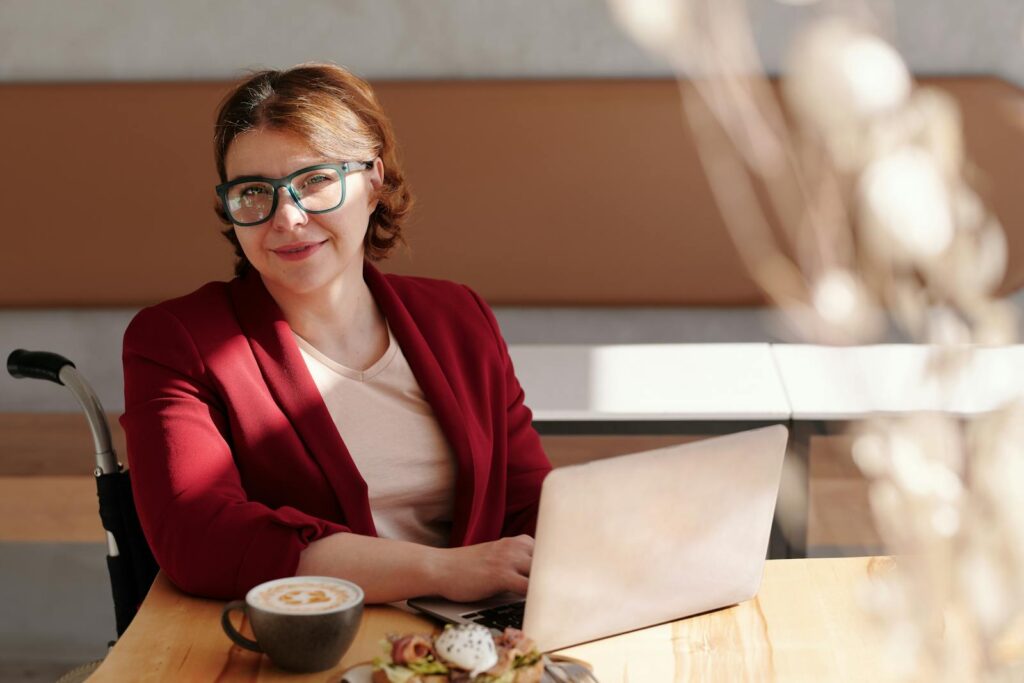 Vagas Remotas na Spun Mídia Smiling woman in red blazer using a laptop at a café table, enjoying her workday.