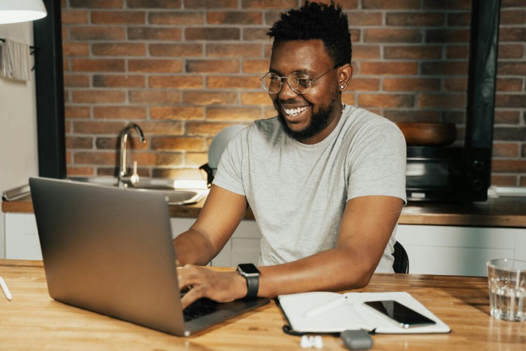 Vagas Remotas na Questor Sistemas African American man smiling while working remotely on laptop from home office