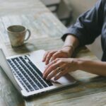Auxiliar de Reservas Remoto e mais Vagas Efetivas em Home Office Crop faceless female freelancer in casual outfit sitting at table with cup of drink and surfing on laptop in light workspace