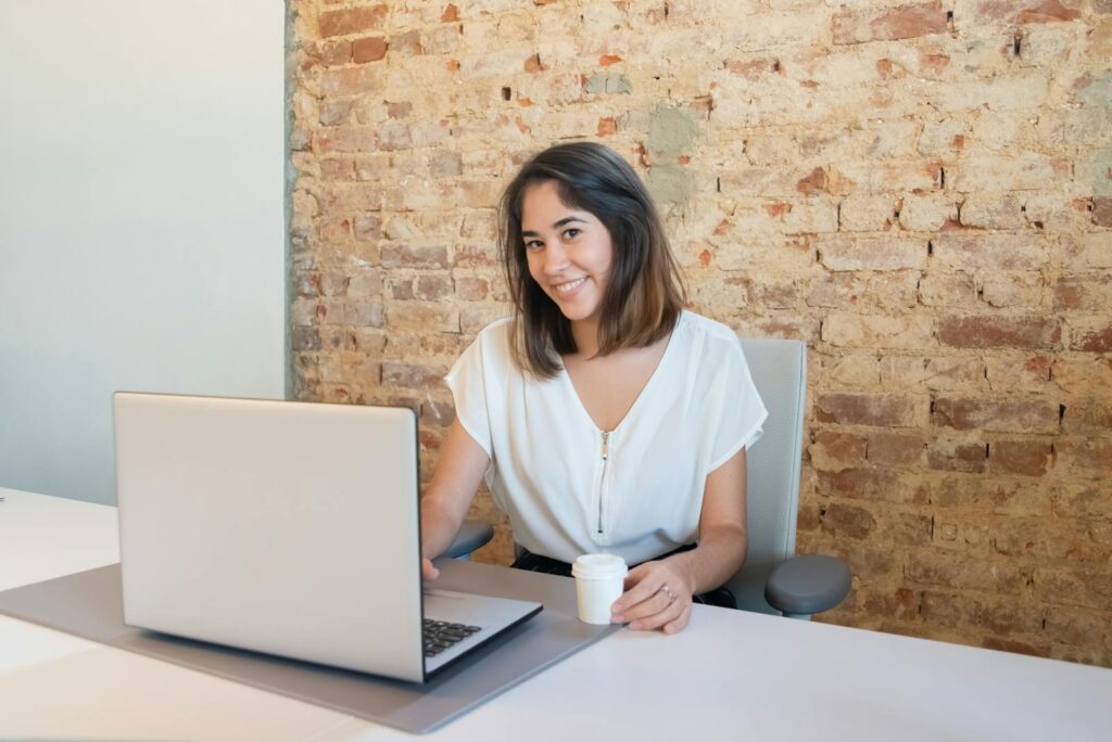 Vagas Remotas na ION Sistemas A cheerful woman working on a laptop in a rustic office setting, holding a cup.