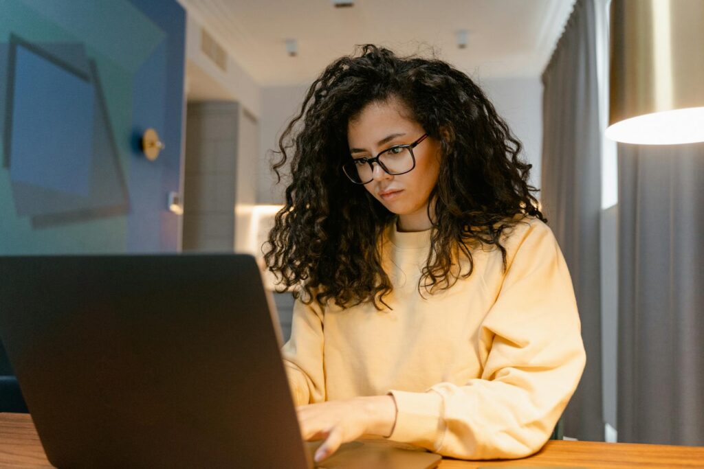 Vagas Remotas na Kedu Woman in yellow sweater working on a laptop in a cozy home setting.