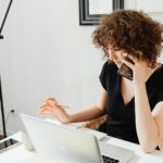 Vagas Remotas na Green4T Woman sitting at table, talking on phone while working on laptop in a home office setting.