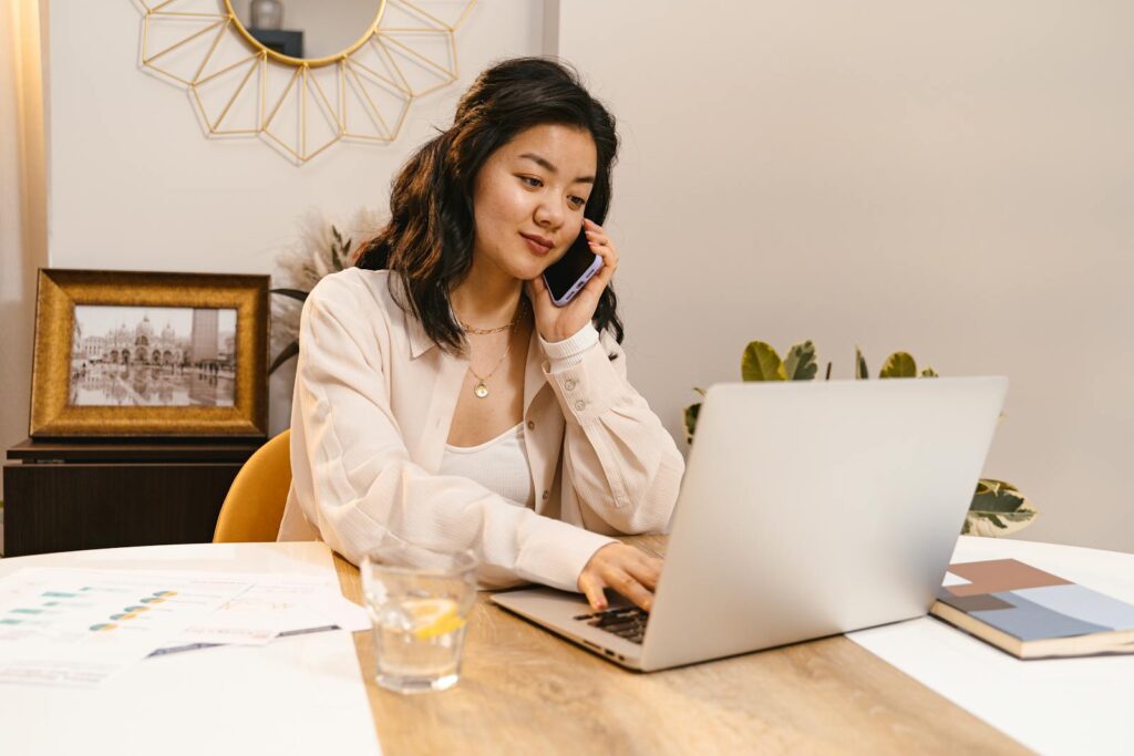 Vagas Remotas na Raffinato Young woman multitasking with laptop and cellphone in home office setup.