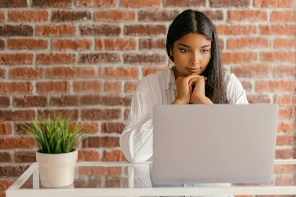 Vagas Remotas para Assistentes em diferentes áreas Young woman deeply focused on her laptop studying indoors against a brick wall backdrop.