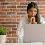 Vagas Remotas para Assistentes em diferentes áreas Young woman deeply focused on her laptop studying indoors against a brick wall backdrop.