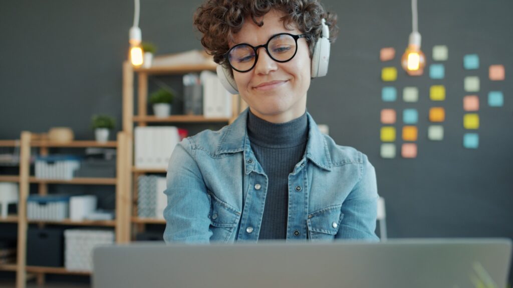 Analista de Suporte Técnico JR e mais Vagas Remotas Abertas! Woman wearing headphones smiles while looking at laptop.