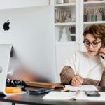 Vagas de Emprego na Beep Saúde A businesswoman multitasks on the phone while writing notes at her home office desk, showcasing modern productivity.