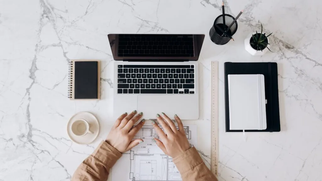 Vagas para teletrabalho na Nexdom Top view of a stylish home office desk with a laptop, planner, and coffee cup, showing hands on a blueprint.