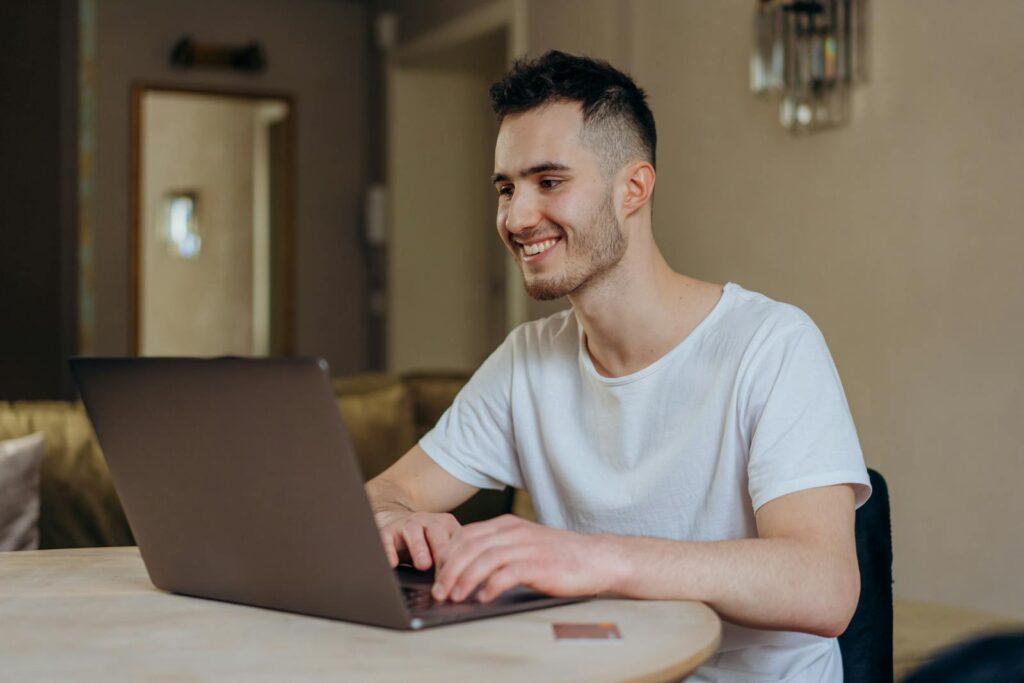 Suporte Técnico JR e mais Vagas Remotas Abertas! Young man smiling while online shopping with laptop indoors at home.