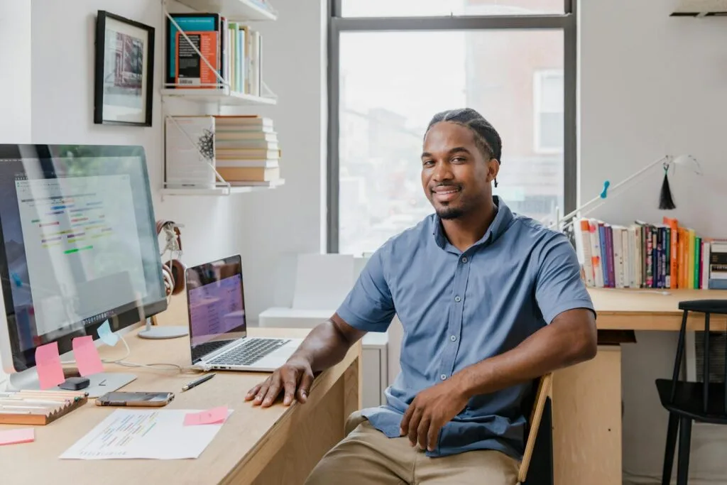 Vagas Remotas no Sicoob, com VA/VR de até R$ 2.300 Confident professional man with a welcoming smile sitting at a desk in a contemporary office setting.