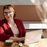 Remotas na Kipiai com benefício flexível de R$ 1.000! Smiling woman in red blazer using a laptop at a café table, enjoying her workday.