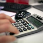 Assistente Fiscal e + vagas (CLT), com R$700,00 no Cartão Caju Close-up of a hand using a calculator on an office desk for accurate calculations.