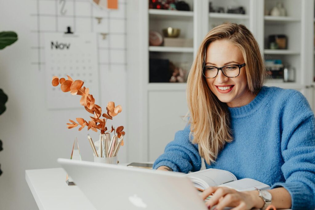 Home Office na Teleperformance, incluindo Aprendiz A woman in a bright sweater smiles while working on her laptop at home.
