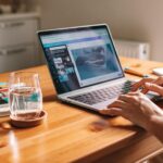Vagas Remotas com VA/VR de até R$ 1.100,00 Close-up of hands on a laptop in a cozy home office with a water glass on desk.
