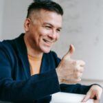 Trabalho Remoto com salário até R$ 6.200 (CLT) Smiling man giving thumbs up while working on laptop from home office, illustrating positive remote work experience.