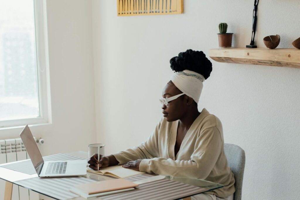 Vagas Remotas na Asaas, VA/VR de R$ 750,00 African American woman writing in a journal while working from home with a laptop.