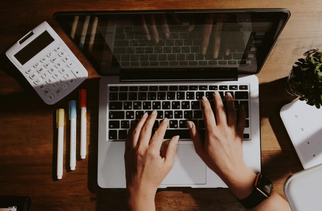 Vagas Remotas na NSTech, VA/VR de R$ 750,00 Hands typing on a laptop with a calculator and pens nearby on a wooden table.