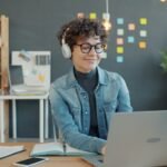 Trabalho Remoto para Atendimento com VA de R$ 1.000,00 Young woman wearing headphones works on laptop in office.