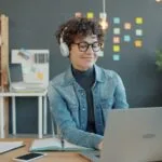 Trabalho Remoto para Atendimento com VA de R$ 1.000,00 Young woman wearing headphones works on laptop in office.