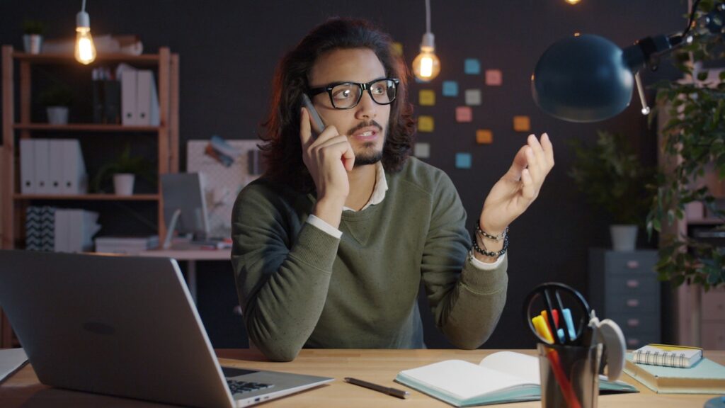 Teletrabalho no Milvus com VA/VR de R$ 900,00 Man talking on phone at desk with laptop.