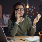Teletrabalho no Milvus com VA/VR de R$ 900,00 Man talking on phone at desk with laptop.